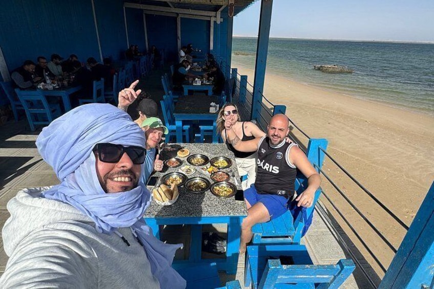 En restaurang vid stranden med blå stolar och bord, en sandstrand och havet som bakgrund. 3