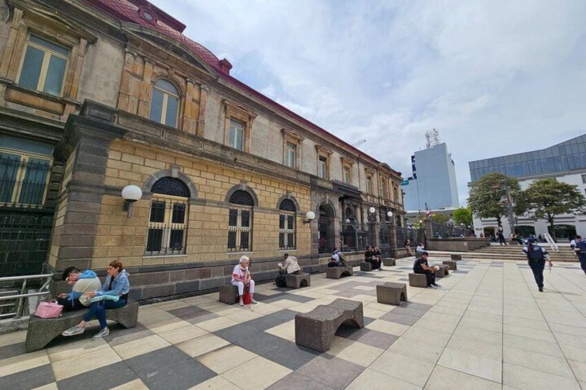 A historic building with arched windows and a red roof, a tiled plaza with benches, and a modern building in the background. 2