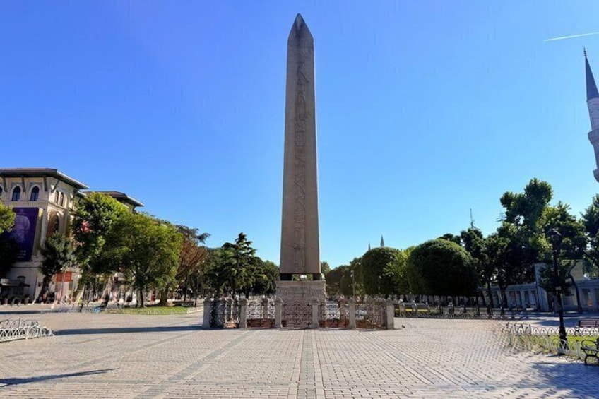 Un alto monumento obelisco en una plaza con suelo empedrado, rodeado de árboles y edificios. 3