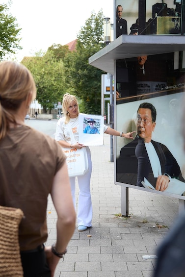 A person holding a sign with a photo and text, standing in front of a glass display case with a portrait inside. 1