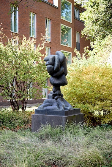 A sculpture of a figure in a contemplative pose, surrounded by grass and trees, with a brick building in the background. 4