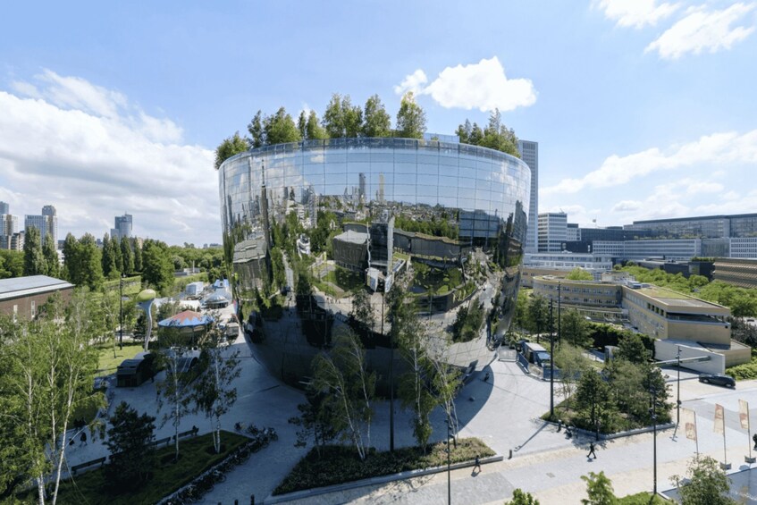 A modern glass building reflecting its surroundings, surrounded by trees and urban buildings. 1