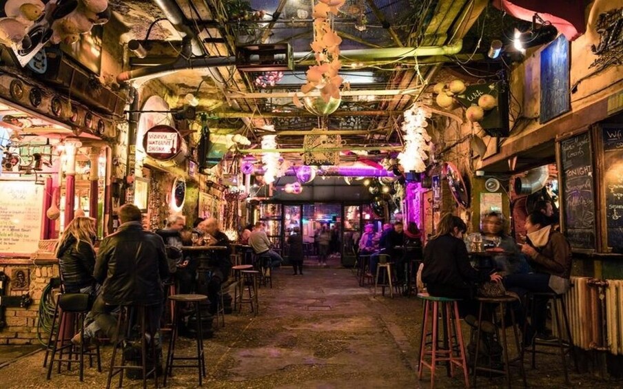 A crowded indoor bar with people sitting at tables, a neon sign, and various decorations on the ceiling. 3