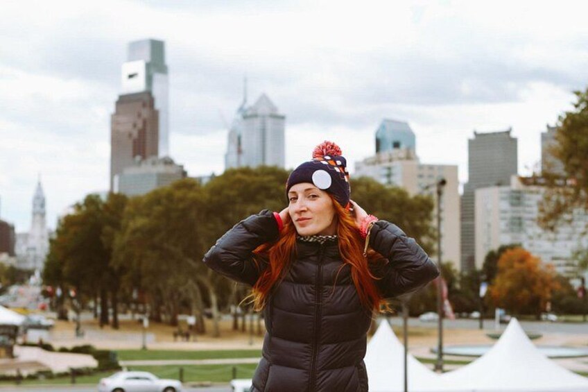 Una persona con chaqueta y sombrero negro, de pie al aire libre con un paisaje urbano de fondo. 1