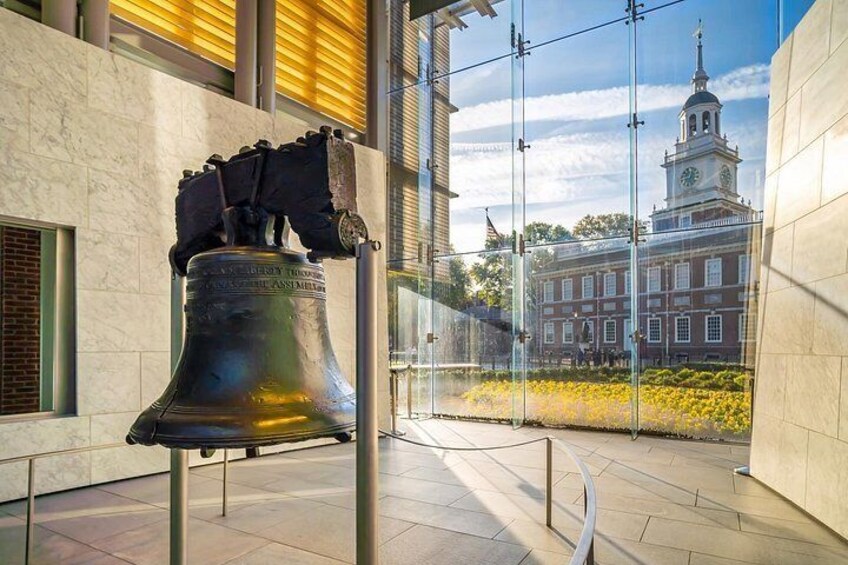 A large bell inside a building with a view of a historic building with a clock tower in the background. 5