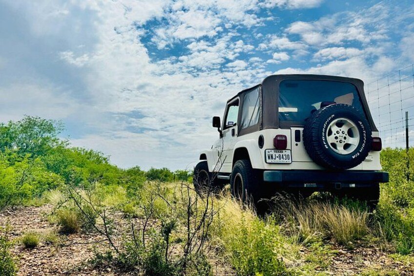 A white off-road vehicle with a spare tire mounted on the back, parked in a grassy field with sparse vegetation and a fence in the background. 2