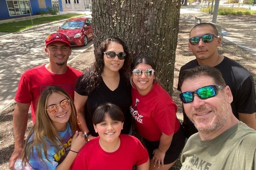 A group of people posing in front of a tree with a red car in the background. 1