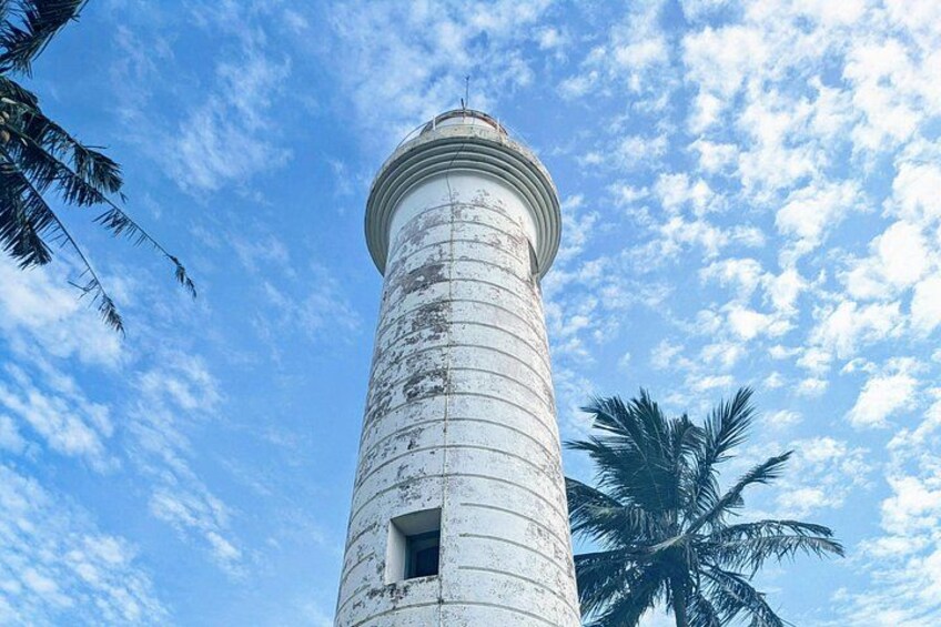 A tall, cylindrical lighthouse with a weathered exterior, set against a backdrop of a clear blue sky with scattered clouds and a palm tree. 2