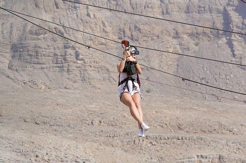 A person suspended in mid-air, wearing a harness and holding a camera, with a mountainous landscape in the background. 3