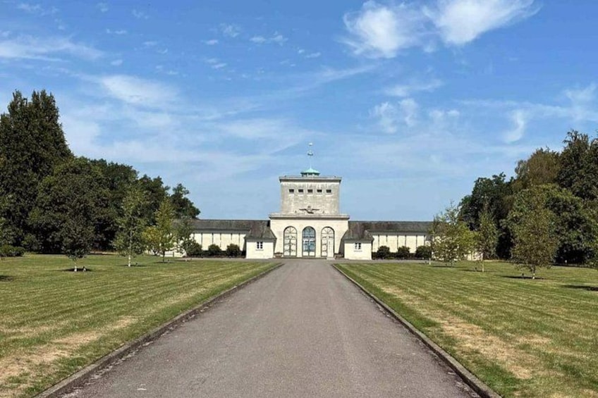 A long driveway leading to a large building with a clock tower, surrounded by well-maintained grass and trees. 1