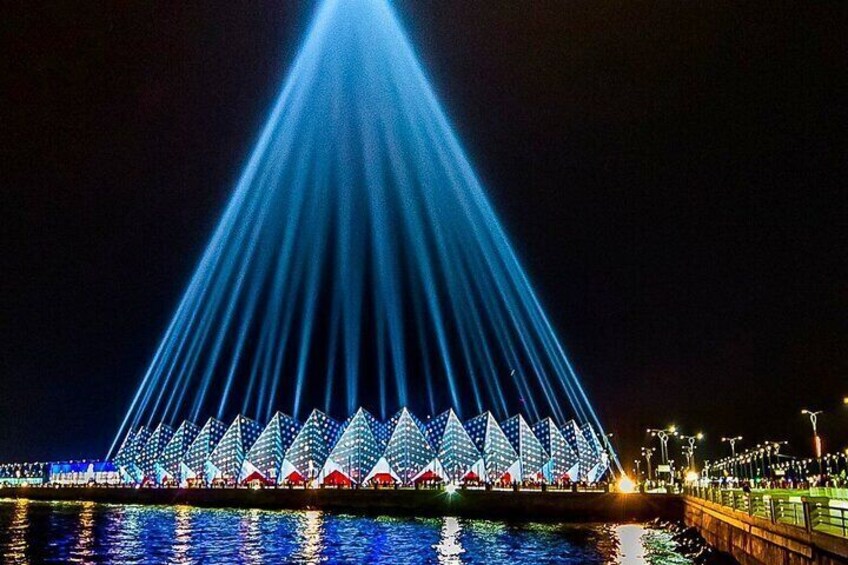 A large illuminated structure with a triangular roof and rows of lights reflecting on water. 1