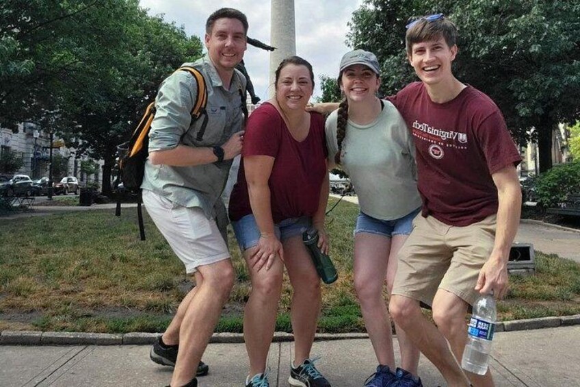 Four people standing on a sidewalk with grass and trees in the background. 3