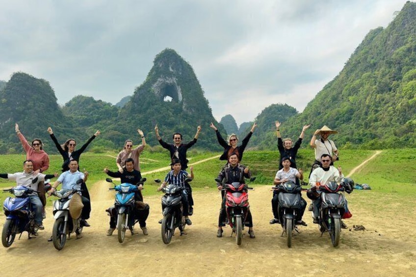 A group of people on motorcycles with their arms raised, on a dirt road with mountains in the background. 2