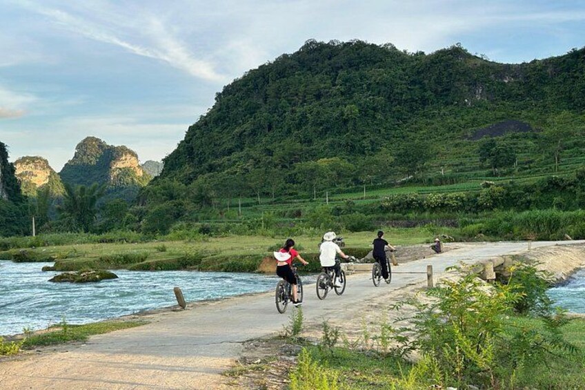 A riverbank with cyclists, a forested hill, and a rocky outcrop in the background. 2