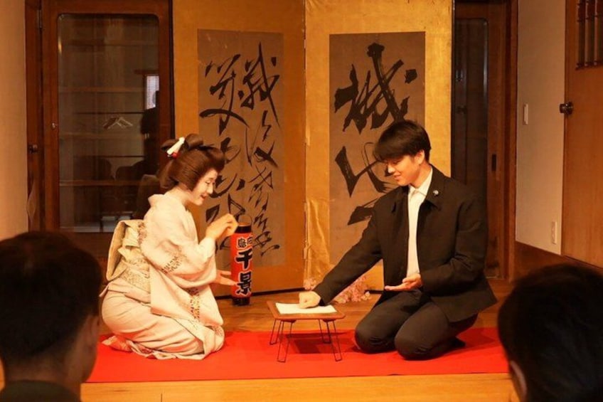 Two people performing a traditional tea ceremony on a tatami mat with calligraphy in the background. 3