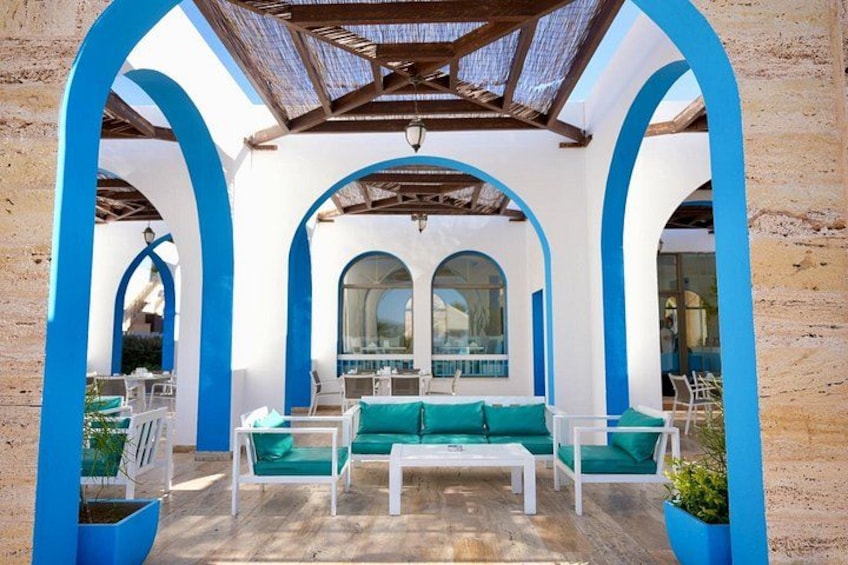 An outdoor seating area with white tables and green cushions, framed by blue arches and a wooden ceiling. 1