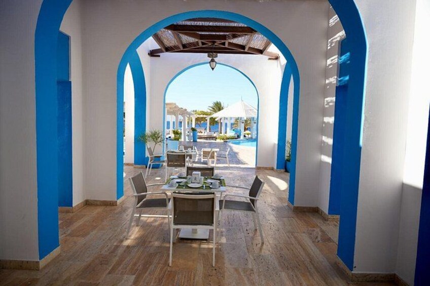 An outdoor dining area with a table and chairs, blue arches, and a view of a pool and buildings in the background. 4