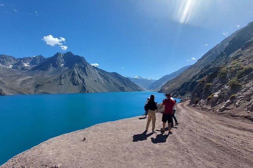 A group of people walking on a dirt path beside a blue lake with mountains in the background. 1