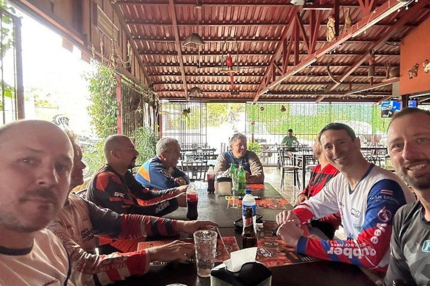 A group of people seated at a table with drinks and a menu, in an outdoor setting with a wooden roof and greenery in the background. 3