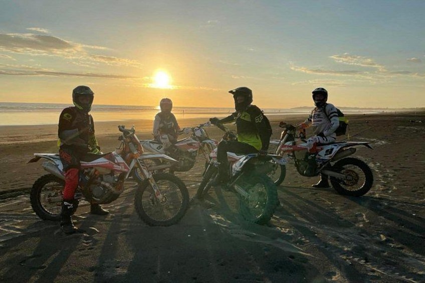 Three motorcyclists on a beach with their bikes, the sun setting in the background. 5