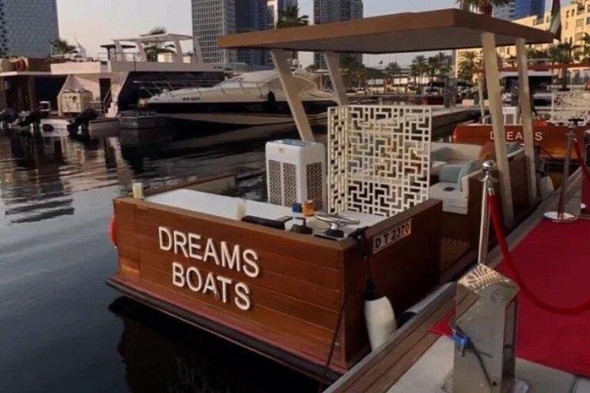 A wooden boat with 'DREAMS BOATS' written on it, docked at a marina with other boats and buildings in the background. 2