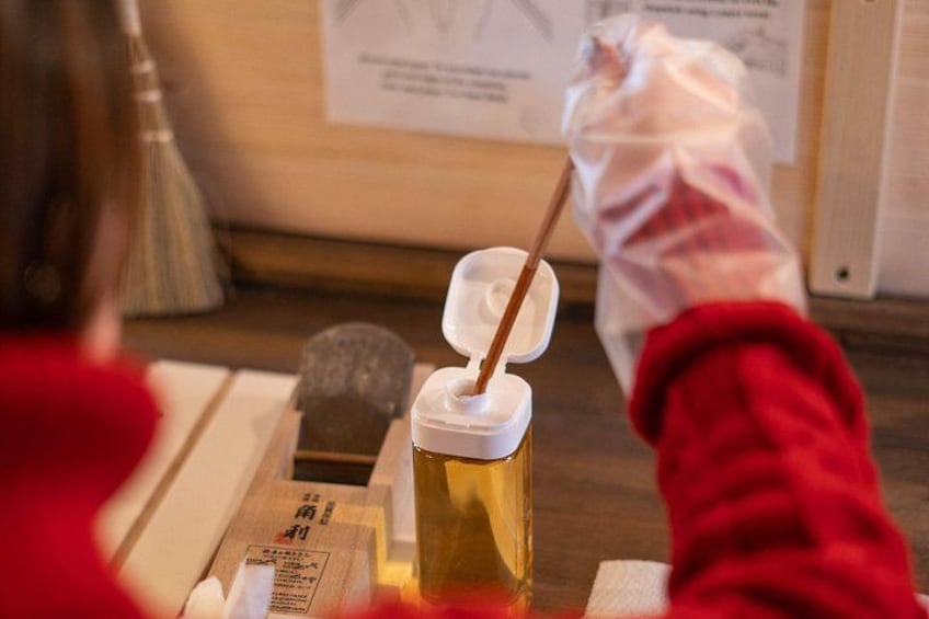 Eine Person mit rotem Pullover und weißen Handschuhen hält eine Flasche mit weißem Verschluss und einem Holzstab. 3