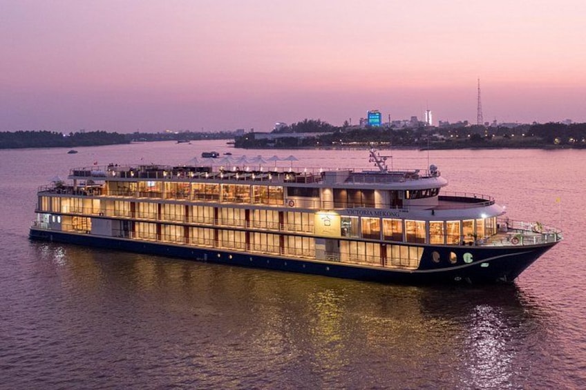 Een groot rivierschip met meerdere dekken en een opvallende boeg, afgemeerd aan een kade met een stadsachtergrond. 1