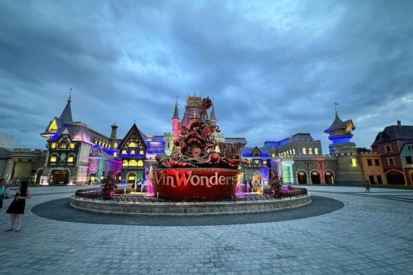 A themed park entrance with a large illuminated sign, a decorative fountain, and castle-like buildings. 2
