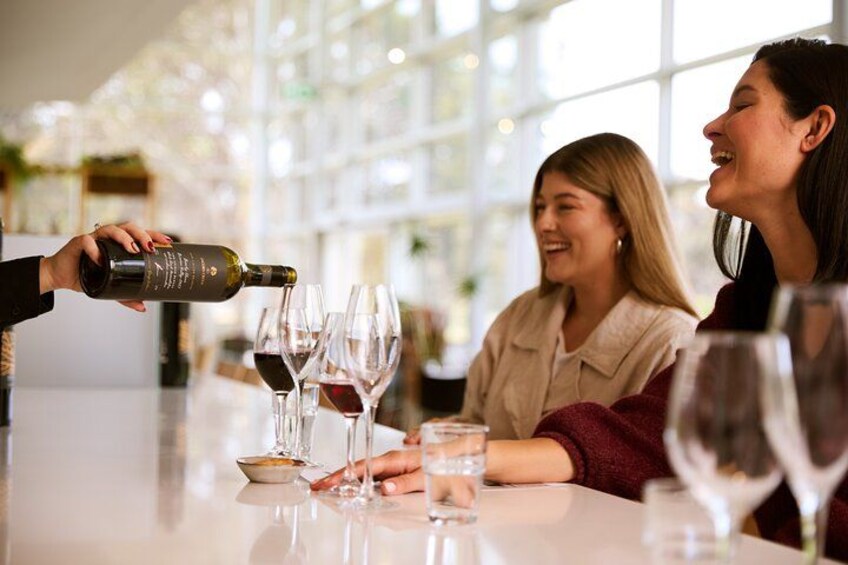 A person pouring wine into glasses on a table in a well-lit room with large windows. 2
