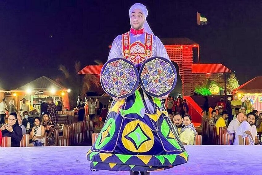 A person in traditional attire with a colorful skirt and a headpiece, standing in front of a crowd at an outdoor event. 2