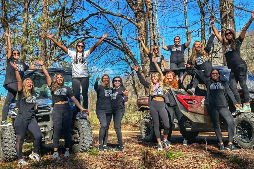 A group of people in black t-shirts posing around an off-road vehicle in a wooded area. 3