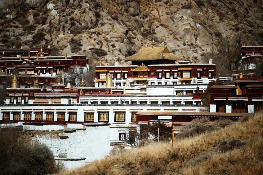 A traditional Tibetan monastery with multiple buildings, a rocky hillside in the background, and a small body of water in the foreground. 2