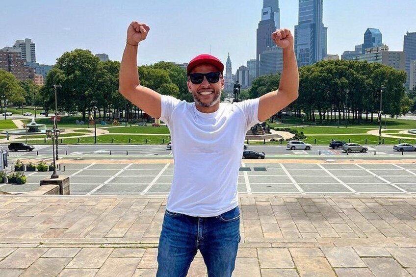 A person standing in a parking lot with raised arms, a city skyline in the background, and a statue in the distance. 5