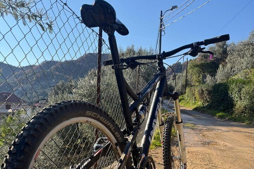 A mountain bike parked against a chain-link fence with a dirt road and hills in the background. 2