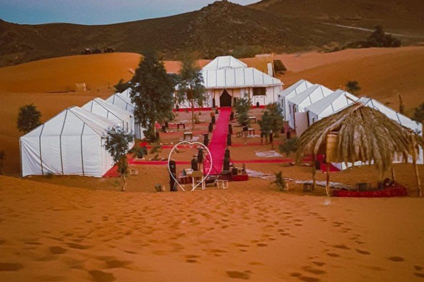 A desert campsite with tents, a red carpet, and a thatched structure. 1