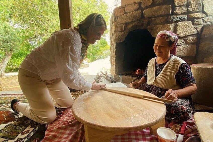 Two people kneeling on a checkered floor, one using a wooden board and the other holding a bowl, in front of a stone oven. 2