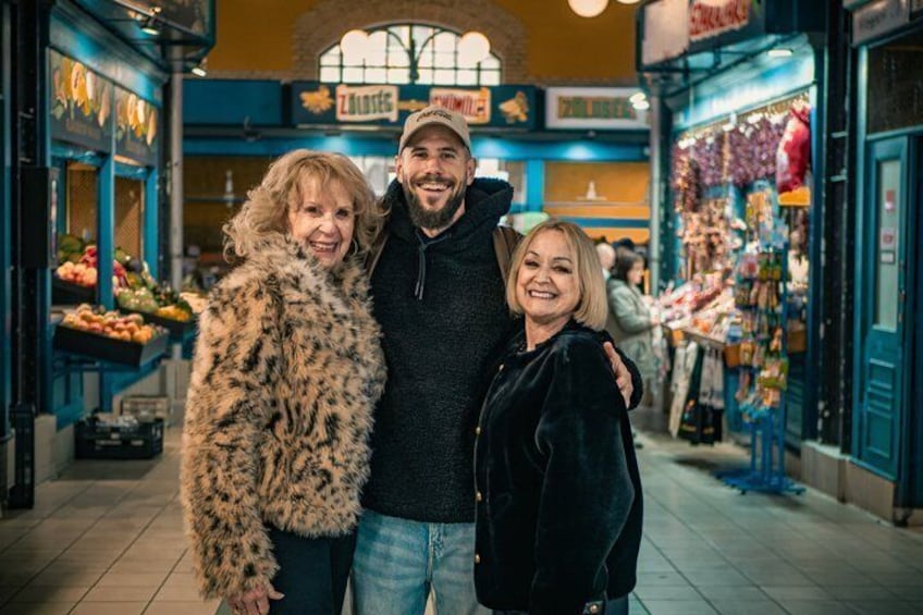 Three people standing in a market with various shops and produce stalls in the background. 2