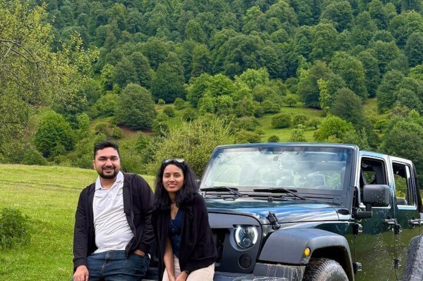 A couple sitting by a black off-road vehicle in a green, hilly landscape. 5