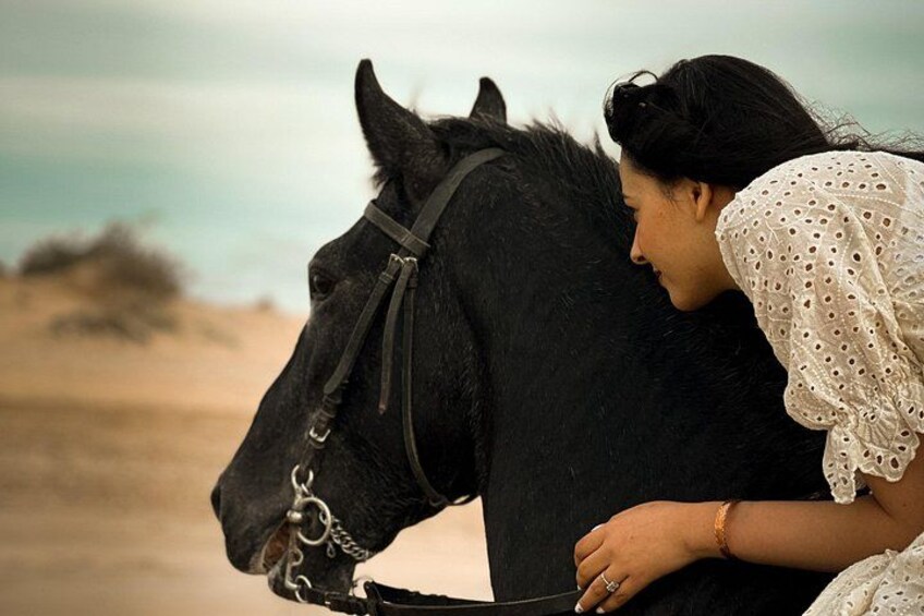 A person leaning on a black horse with a bridle, wearing a white blouse with lace details, in a desert-like setting with sandy ground and sparse vegetation. 2