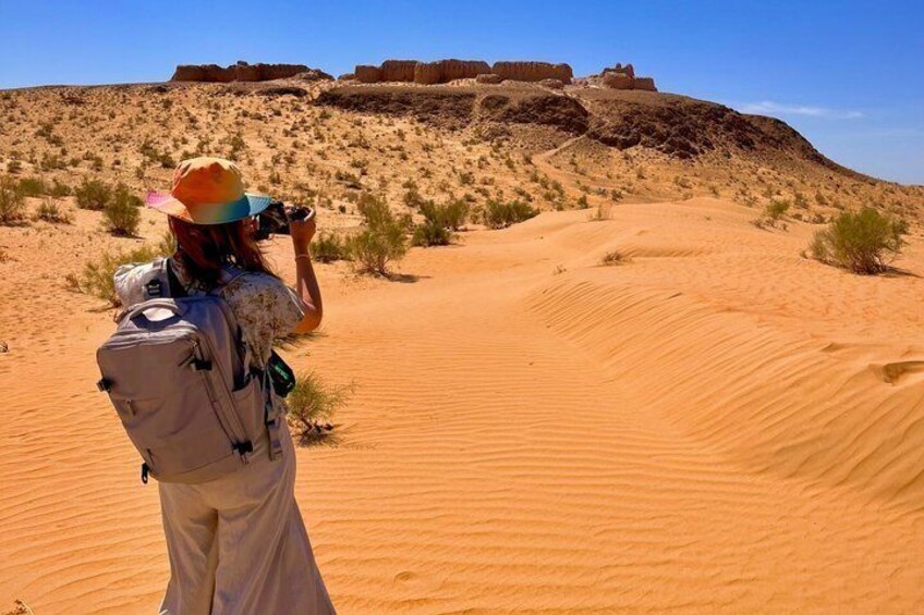 A person taking a photo of a desert landscape with sand dunes and a distant fortification. 3