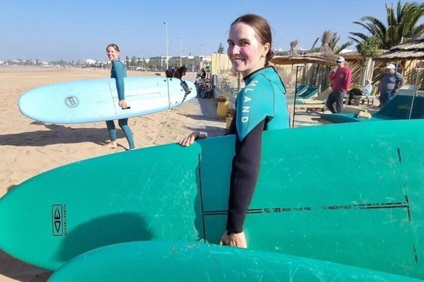 Una persona sostiene una tabla de surf en la playa, con palmeras y otras personas al fondo. 2