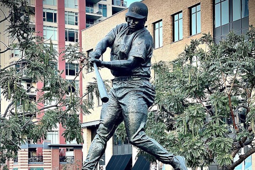 En bronzestatue af en baseballspiller i svingposition, omgivet af træer og bygninger. 4