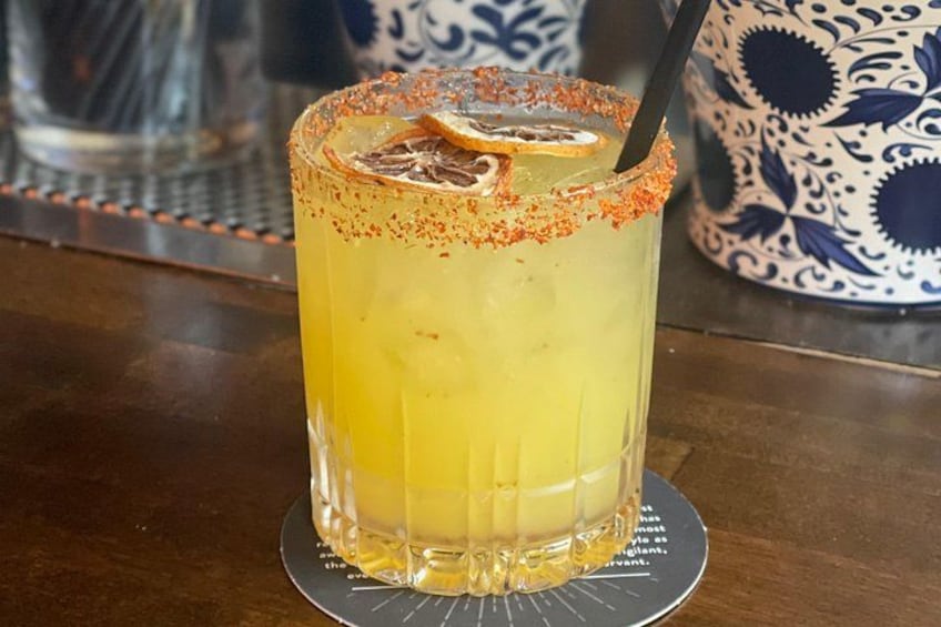 A tall glass of yellow beverage with a rim of red seasoning, a slice of orange, and a straw. 2