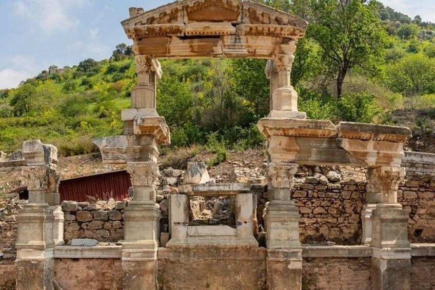Ruinas antiguas de piedra con columnas y un arco parcialmente conservado, frente a un paisaje de colinas verdes. 4