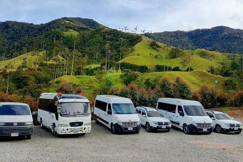 A row of white vans parked on a gravel surface with a hilly landscape in the background. 1
