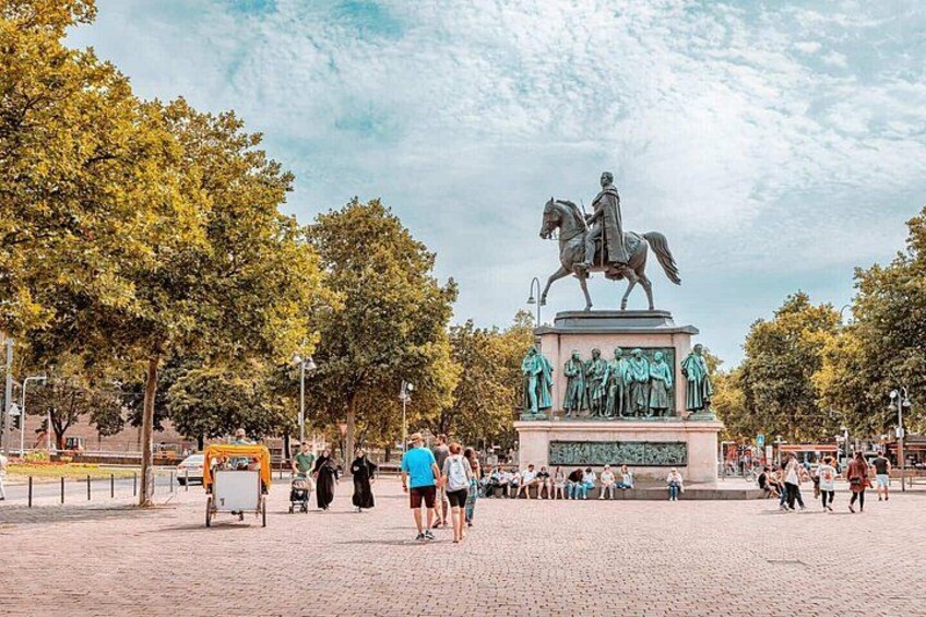 Una statua raffigura un uomo a cavallo, circondato da diverse figure, in una piazza pubblica con alberi e persone. 3