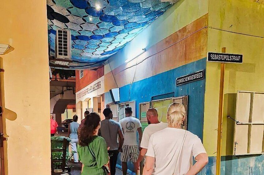 Un corridoio colorato con un soffitto decorato, un segnale con la scritta 'CONSCIENTIESTEG' e una porta con l'indicazione 'SEBASTOPOLSTRAAT'. 2