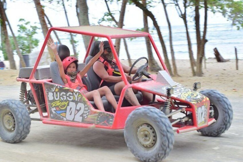 A red buggy with two passengers, one child and one adult, on a sandy beach with trees and the ocean in the background. 3
