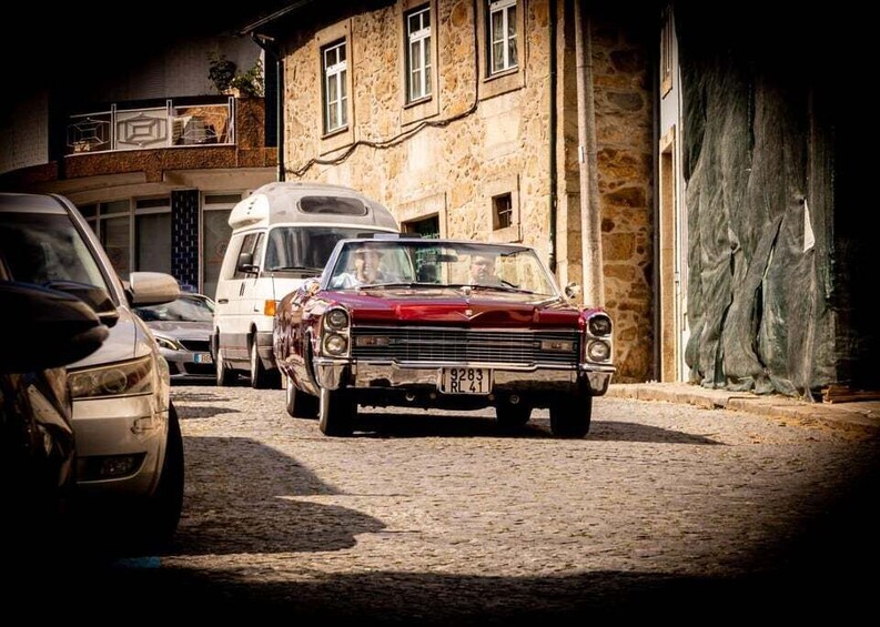 A vintage red car parked on a cobblestone street, a white van, and a stone building with a balcony. 1