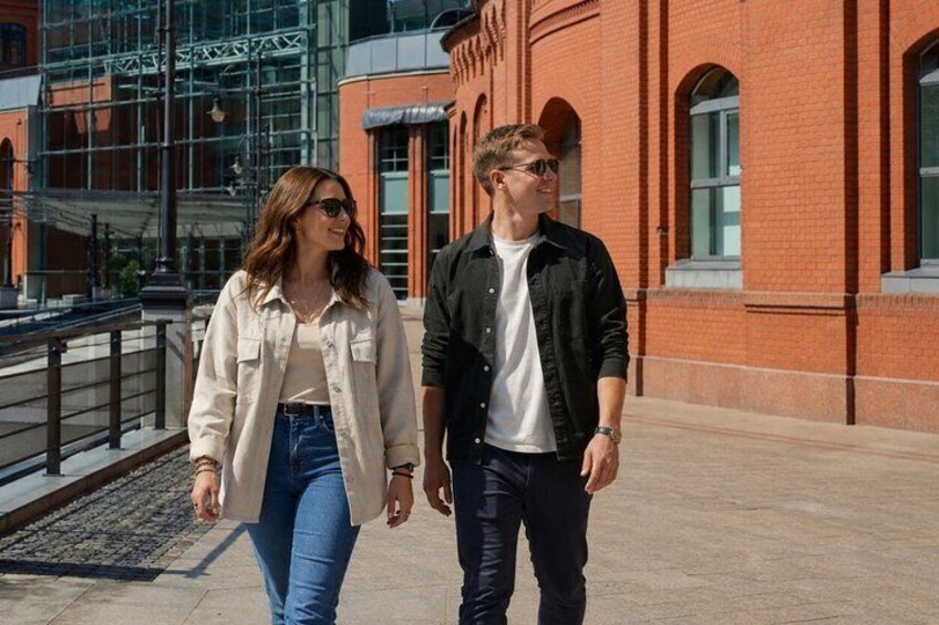 Two people walking on a sidewalk in front of a red brick building with large windows and a metal structure in the background. 1